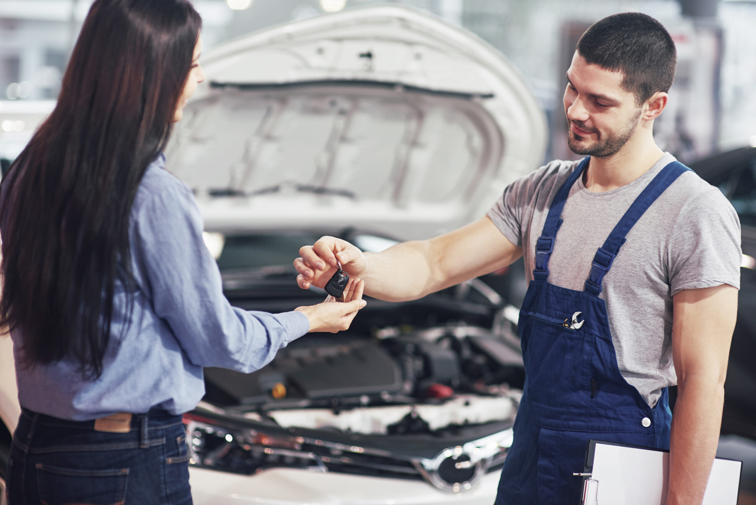 A mechanic in overalls hands car keys to a woman standing in front of a white car with its hood open.